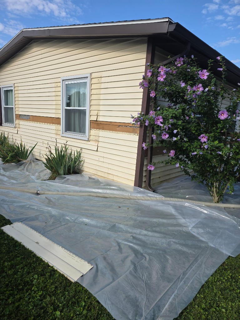 Cream-sided home with siding removed, insulation hose on the ground, and plastic sheeting protecting the lawn
